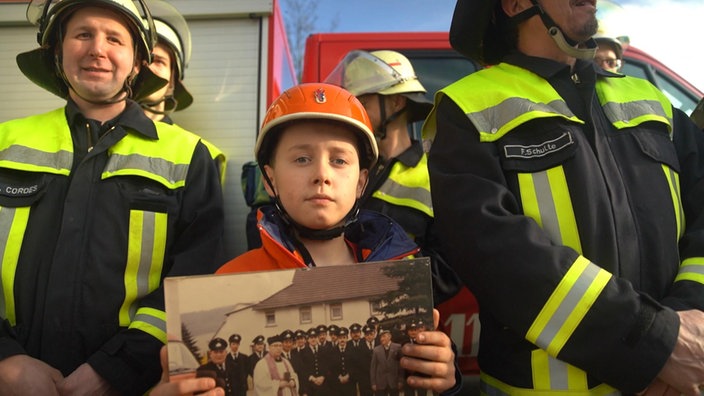 Ein Kind in Feuerwehrkleidung hält ein altes Feuerwehrbild in der Hand.