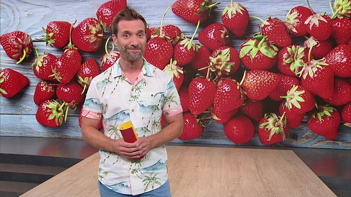 Daniel Aßmann steht im Servicezeit-Studio. Hinter ihm ist ein Bild von losen Erdbeeren zu sehen.