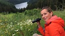 Tamina Kallert mit Fernglas schaut von einer Höhe auf einen darunter liegenden Waldsee