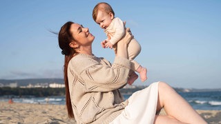 Lächelnde Frau am Strand hält ihr Kind in die Höhe. 