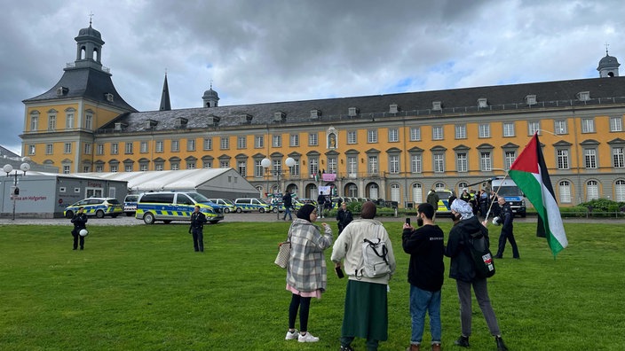 Menschen mit Palästina-Fahne vor dem Hauptgebäude der Uni in Bonn. Vor dem Gebäude stehen zahlreiche Polizei-Wagen