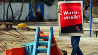 Archivbild. Ein Mensch hält ein Schild mit der Aufschrift "Warnstreik" auf dem Spielplatz einer Kindertagesstätte