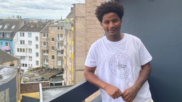 Ein dunkelhäutiger junger Mann steht in einem weißen T-Shirt vor einer schwarzen Wand auf einem Balkon und lächelt