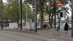 Auf dem Foto ist der Worringer Platz in Düsseldorf. In der Mitte der Platzes sitzen Männer in schwarzen Hoodies auf einer Bank.