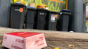 Eine Verpackung liegt auf der Straße vor mehreren vollen Mülltonnen.