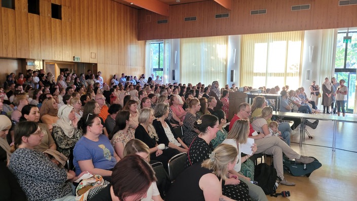 Viele Menschen in Stuhlreihen in einem vollen Saal, vorne spricht eine Frau mit Mikrofon