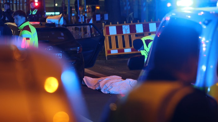  Eine tote Person liegt abgedeckt neben einem Fahrzeug mit zersplittertem Fenster unter einem Tuch.