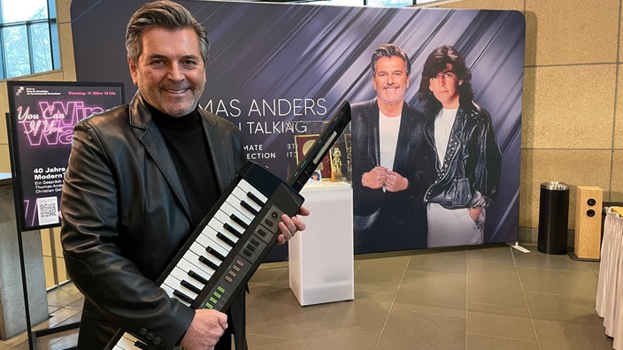 Thomas Anders mit einem Umhängepiano in der Hand vor einem großen Banner und einer Vitrine mit Ausstellungsstücken