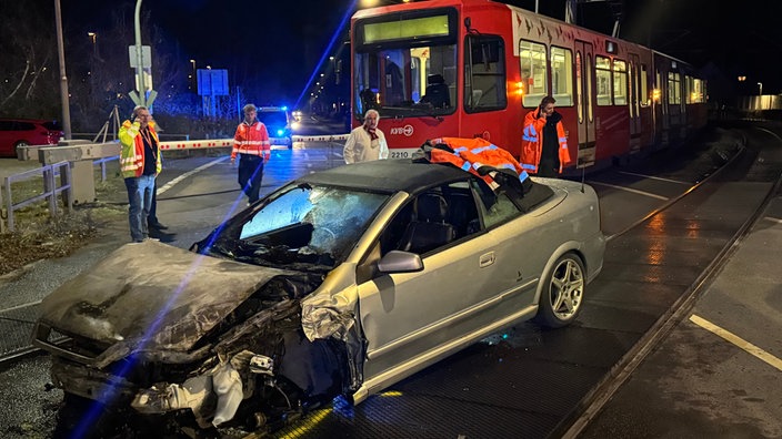 Silbernes Cabrio mit demoliertem Frontbereich, im Hintergrund Einsatzkräfte und Straßenbahn