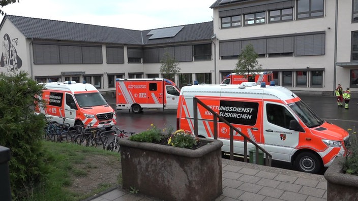 Drei Rettungswagen stehen neben Fahrradständern und vor einer Treppe auf einem Schulhof.