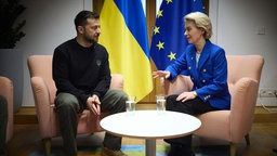 Ursula von der Leyen und Wolodymyr Selenskyi sitzen an einem Tisch und reden. Im Hintergrund die EU-Flagge, sowie die ukrainische Flagge.