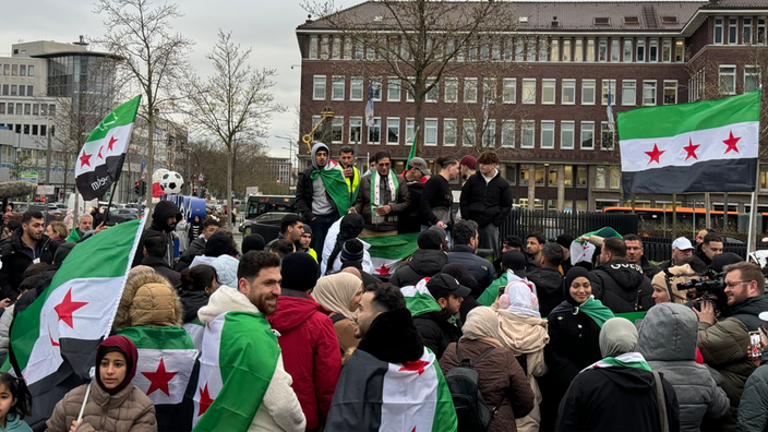 Mehrere Leute stehen im Halbkreis um Menschen auf einer Empore und halten Flaggen