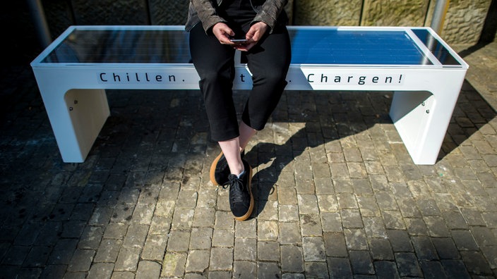 Auf dem Foto ist eine Bank ohne Lehne, die Sitzfläche ist eine verglaste Solarzelle. Auf der Bank steht "Chillen." und "Chargen!".