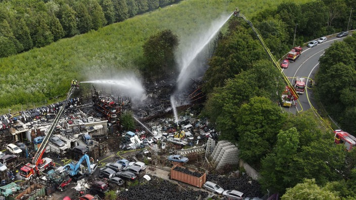 Viele Autos auf einem Schrottplatz, darum Feuerwehr-Fahrzeuge mit Drehleitern.