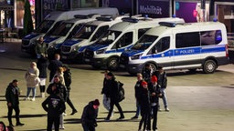 Menschen stehen auf einem Platz. Im Hintergrund stehen Mannschaftswagen der Polizei.