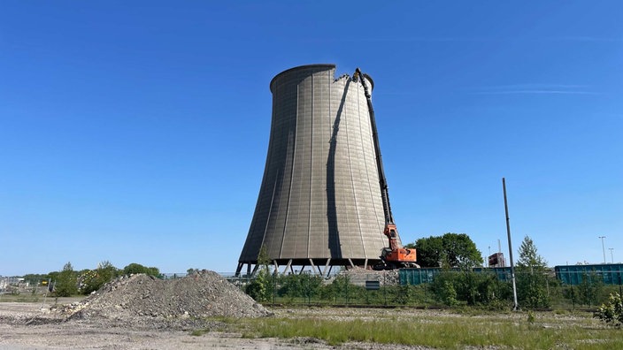 Ein langarmiger Bagger beginnt von oben mit dem Abriss eines alleinstehenden Kühlturms. Blauer Himmel