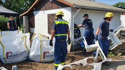 Männer in blauen Uniformen vor einer Gartenlaube umgeben von weißen Säcken mit Erde