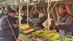 Ein älterer Herr steht vor einem Marktstand und schaut auf den Marktbeschicker, der ihm ein paar Sachen einpackt