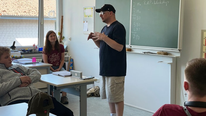 Eine Mann steht im Klassenraum vor einer Tafel und spricht, drei Zuhörer sitzen drumherum.