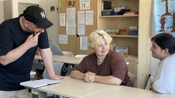 Marko Cebi steht in einer Schulklasse an einem Tisch und schaut in ein Notizbuch. Die Schülerinnen Selina und Felicitas sitzen daneben und schauen sich an.