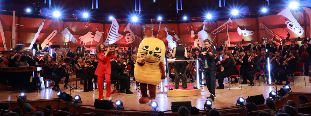 Die Maus, Jana Forkel und Johannes Büchs stehen gemeinsam mit Musiker*innen des WDR Sinfonieorchesters auf der Bühne der Kölner Philharmonie.