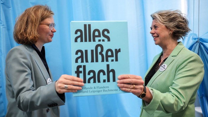 Die Kuratorinnen Bettina Baltschev (l) und Margot Dijkgraaf stehen mit dem Motto des Gastlandes 2024 ·alles außer flach· auf der Leipziger Buchmesse. Die Niederlande und Flandern wollen sich als Gastland auf der Leipziger Buchmesse 2024 präsentieren.