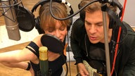 Mann und Frau mit Kopfhörern im Studio vor Mikros am singen, er spielt zusätzlich Gitarre.