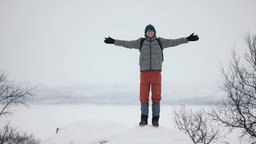 Ein glücklicher Mann in warmen Kleidern steht mit ausgestreckten Armen auf verschneitem Terrain in Finnland.