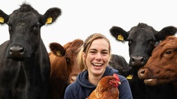 Marie vom Tiggeshof hält ein Huhn in der Hand und lächelt in die Kamera. Im Hintergrund sind Kühe zu sehen