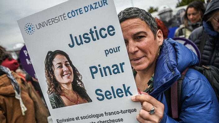 Das Beitragsbild des Dok5  "Pinar Selek – türkische Soziologin und Menschenrechtlerin" zeigt eine Demonstrantin vor dem Gerichtsgebäude in Istanbul 2023 mit einem Protestschild mit der Aufschrift Gerechtigkeit für Pinar Selek.  