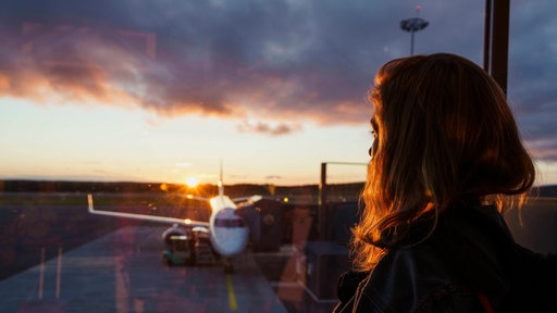 Junge Frau blickt bei Sonnenuntergang aus Flughafenfenster auf Flugzeug auf dem Rollfeld