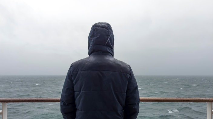 Ein Mann steht mit Jacke an der Rehling eines Kreuzfahrtschiffes und schaut bei Regen auf das Meer hinaus