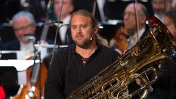 Andreas Martin Hofmeir mit seiner Tuba vor einem Orchester in Nahaufnahme.