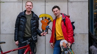 Daniel Schlipf (links) und André Gatzke vor einer Wand, auf der ein Graffiti der WDR-Maus zu sehen ist. 