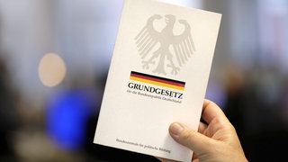 Nahaufnahme einer Ausgabe des Grundgesetzes, die von der Hand einer Person ins Bild gehalten wird. 