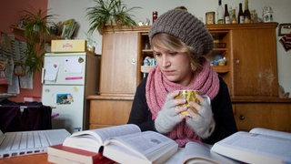 Ein Studentin sitzt mit Mütze und Handschuhen am Tisch und lernt.