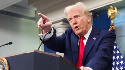 Donald Trump – ein alter Mann mit weißem Haar, blauem Anzug und roter Krawatte – steht an einem Rednerpult und zeigt mit dem Finger auf jemanden oder etwas außerhalb des Bildes.