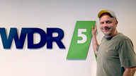 Ralph Ruthe posiert vorm WDR5 Logo