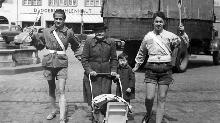 Zwei Schülerlotsen haben eine Frau mit Kinderwagen und einem kleinen Jungen in die Mitte genommen und führen sie mit erhobenen Kellen über eine Straße (Aufahme von 1953)