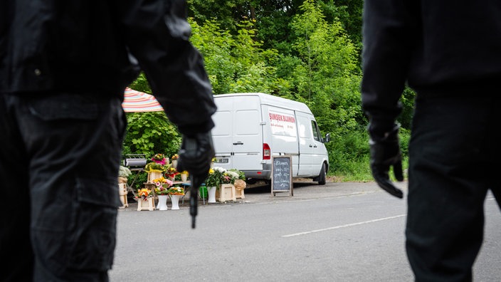 Zwei bewaffnete Männer von hinten schauen auf einen weißen Kleintransporter mit Blumenhandel