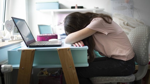 Ein Mädchen sitzt am Schreibtisch, vor sich einen Laptop, und hat den Kopf auf die Arme gelegt.