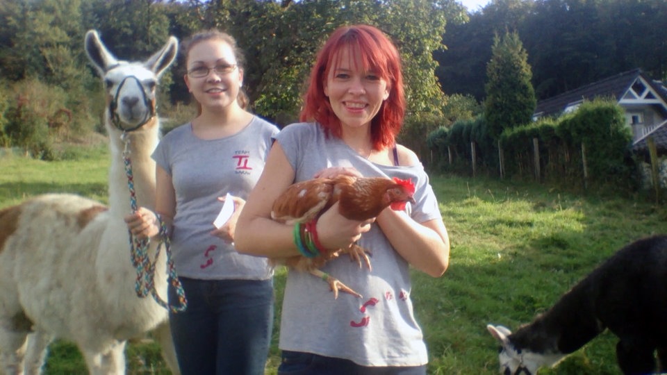 Zwei Frauen lachen in die Kamera, eine hält ein Lama, die andere ein Huhn