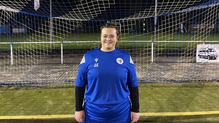 Frau im blauen Fußballtrikot steht vor einem Tornetz, lächelt frontal in die Kamera. Der Hintergrund zeigt das Stadion und das Spielfeld.