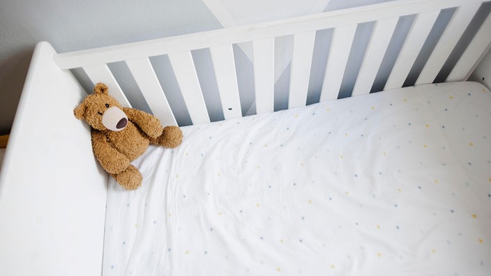 Das Bild zeigt ein leeres weißes Babybett mit einer weißen Matratze und einem braunen Teddybären in der linken Ecke.