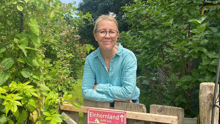 Frau lehnt sich über das Tor eines Gartens. Im Hintergrund sind viele grüne Bäume zu sehen.