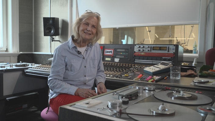 Eine lächelnde Frau mit Brille auf dem Kopf sitzt im Tonstudio vor einem analogen Tonbandgerät.