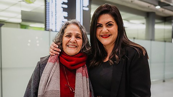 Mutter und Tochter stehen eng beieinander vor einer Anzeigetafel am Flughafen und lächeln freundlich in die Kamera