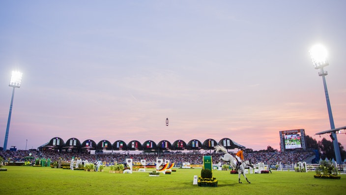 Das Reitstadion in abendlicher Stimmung, im Vordergrund Pferd und Reiter an einem Hindernis, im Hintergrund die Tribüne.