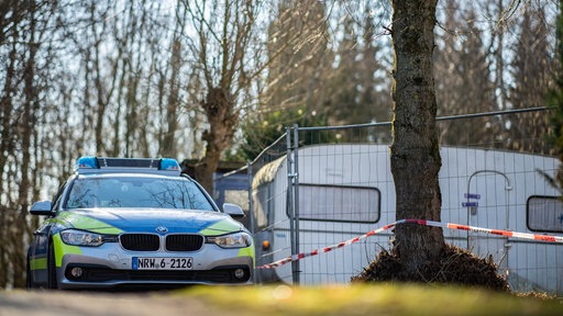 Ein Polizeiauto steht neben einem abgesperrten Grundstück, im Hintergrund ein Campingwagen