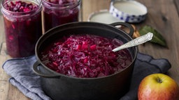 Rotkohl mit Äpeln in einem eisernen Topf, dahinter Rotkohl in Gläsern und im Vordergrund ein Apfel.